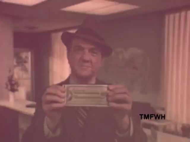 The 16mm still from the 1970s American Express commercial featuring Karl Malden showcases him holding an American Express card. Malden, dressed in a fedora and suit, conveys a sense of earnestness, reinforcing the iconic tagline 'Don't leave home without them.' The setting appears to be an office, indicated by the background decor, which includes a map and some plants, embodying the commercial's mid-century aesthetic. The warm, slightly faded colors evoke the vintage feel of the era.