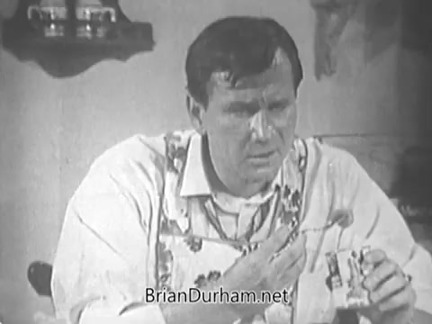 The still from 'March of Dimes Mothers' March - 1960's' features a man wearing an apron, conveying a serious expression while holding a spoon and what appears to be a can, likely involved in a domestic or cooking scenario. The setting hints at a home environment, emphasizing themes of family and caregiving relevant to the Mothers' March campaign promoting health and support for children. The black-and-white format underscores the period's aesthetic.