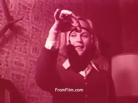 The still from 'Bayer Works Wonders' depicts a woman holding a small object, possibly a medicine or product, while appearing engaged and expressive. The scene is characterized by a warm, reddish tone, which adds to the nostalgic feel typical of early 1970s cinematography. The background features patterned wallpaper, hinting at the era's design trends. The emphasis on the woman's interaction with the object suggests themes of discovery or promotion related to Bayer's products.
