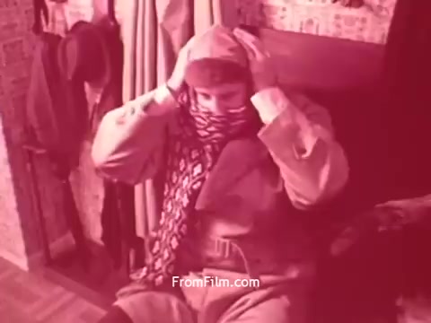 The still from 'Bayer Works Wonders' depicts a man seated indoors, wearing a coat and a scarf over his head. He appears to be adjusting the scarf while looking down. The surroundings are suggestive of a domestic setting, with hanging coats visible in the background. The image has a reddish tint, typical of early colored films, contributing to a vintage aesthetic. The overall mood conveys a sense of preparation or contemplation.