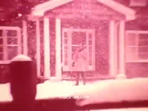 The still from 'Bayer Works Wonders' depicts a winter scene with snowfall in front of a house. A figure in a coat stands near the entrance, surrounded by a thick blanket of snow. The overall color has a reddish tint, typical of 16mm prints from that era, contributing to a nostalgic atmosphere. The focus on the person suggests a moment of pause or contemplation in the snowy landscape.