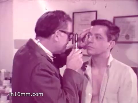 The still depicts a medical examination in a doctor's office. A doctor, wearing glasses and a lab coat, is closely examining a young man's eye with an instrument. The patient, who appears to be shirtless, looks directly at the doctor with a focused expression. The setting is typical of a 1960s medical environment, featuring framed certificates on the wall, suggesting a professional atmosphere. The overall tone reflects the serious health message conveyed in the accompanying voice-over about the importance of flu vaccination.