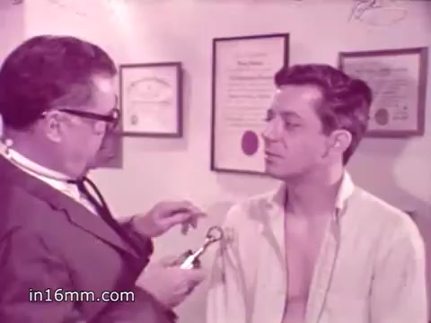 The still shows a doctor preparing to administer a flu vaccine to a young man. The doctor, wearing glasses, holds a syringe as he leans in towards the patient's arm, which is exposed. The young man appears calm but slightly apprehensive. In the background, framed certificates and documents hang on the wall, indicating a medical office setting. The overall tone reflects the era's public health messaging about vaccination and prevention.