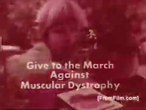 The still features a young child with light hair, appearing in a close-up shot. Bold text overlays the image, reading 'Give to the March Against Muscular Dystrophy.' The overall tone is earnest, likely aiming to encourage support for the cause. The visual quality reflects the 16mm film format, with a warm coloration that contributes to the emotional impact of the public service announcement, which is accompanied by a voice-over from Joan Crawford.