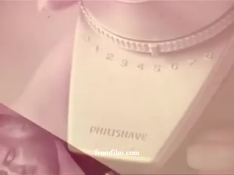 The still features a close-up of a Philishave razor, highlighting the brand name prominently. The design of the razor is retro, showcasing a sleek and simple control with numbered options. The image exhibits a faded, warm color palette typical of 1970s film, giving it a nostalgic feel. In the background, there appears to be a blurred figure, suggesting a human presence. The overall composition combines product advertisement with an intimate, personal touch.