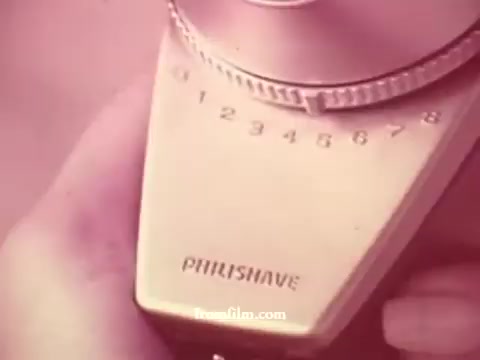 The still features a close-up of a Philishave electric shaver from the 1970s. The device includes a dial with a numeric scale ranging from 1 to 8 and is prominently labeled 'PHILISHAVE.' The overall aesthetic has a vintage, nostalgic vibe, typical of advertisements from that era, often emphasizing innovation in grooming technology. The color palette appears muted, likely reflecting the film's age and the 16mm format.