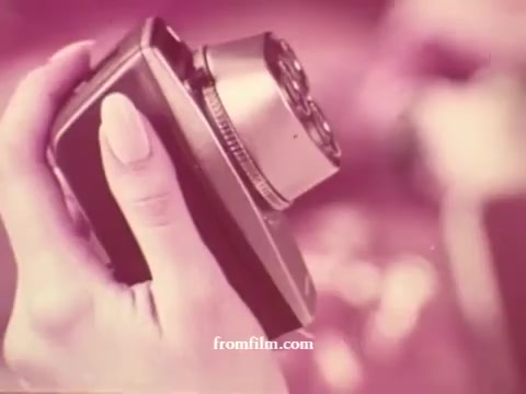 The still features a close-up of a hand holding a compact, vintage camera. The camera has a metallic finish and a circular lens, suggesting a mid-20th century aesthetic. The image is tinted in warm tones, typical of 1970s film stock, creating a nostalgic atmosphere. The background remains blurred, focusing attention on the camera.