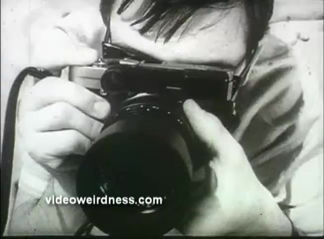 The still from the 'Out of It' trailer features a close-up of a young man holding a camera, preparing to take a photograph. His focused expression and the vintage style of the camera evoke a sense of nostalgia. The black-and-white texture of the 16mm print enhances the retro aesthetic, characteristic of late 1960s cinema. The scene captures a moment of candid engagement, typical of the film's exploration of personal and societal themes.