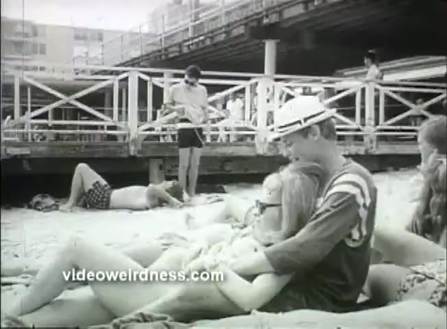 The still from the trailer of 'Out of It' depicts a beach scene featuring a group of young adults. In the foreground, a couple sits close together, with the male character embracing the female, who wears sunglasses. In the background, other beachgoers are seen lounging and socializing, with a pier structure visible above them. The overall tone is relaxed, reflective of the era's youthful spirit, presented in black and white.