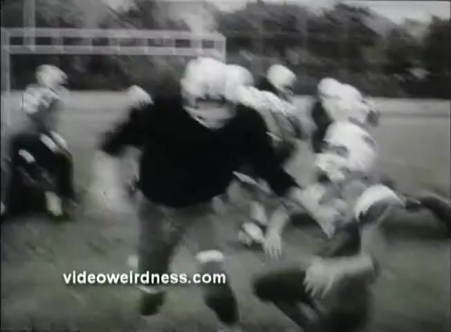 The still from the trailer for 'Out of It' (1969) features a high school football practice scene. In the foreground, a player in a black jersey and helmet appears to be tackling or approaching another player, who is on the ground. Surrounding them are several other players in football gear, indicating a chaotic practice environment. The image is in black and white, typical of the film's era, and captures a moment of physical intensity on the field. The background shows a football field, adding context to the scene.