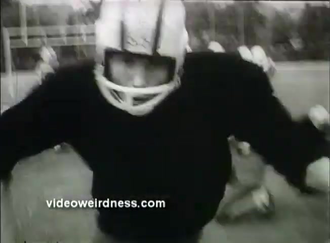 The still from the 'Out of It' trailer features a close-up of a football player wearing a helmet and a black jersey, charging towards the camera. The background shows other players in motion, creating a dynamic and intense atmosphere typical of sports scenes. The image captures a sense of urgency and energy, characteristic of the film's youthful themes.