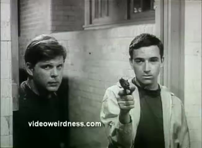 The still from the trailer of 'Out of It' (1969) features two young men in a tense moment. One man, on the left, has a serious expression and is looking off-camera, while the other, on the right, is pointing a gun towards the viewer. The setting appears to be a dimly lit hallway, adding to the suspenseful atmosphere. The overall tone suggests a confrontation or a moment of high tension.