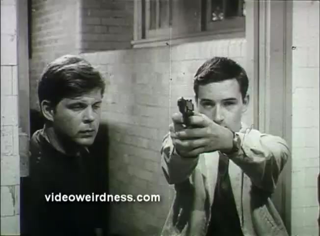 The still from the 1969 trailer for 'Out of It' features two young men in a tense moment. One man, positioned closer to the camera, holds a handgun aimed forward, while the other looks on with a concerned expression. The setting appears to be a hallway with light-colored walls, indicating a potentially dramatic or confrontational scene. The overall tone suggests themes of conflict or confrontation.