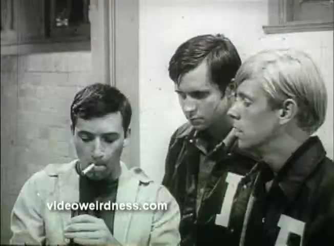 The still from the 1969 trailer for 'Out of It' features three young men in a dimly lit room. One of them, the foreground figure, is smoking a cigarette, appearing thoughtful or contemplative. The other two stand nearby, looking on with interest or concern. They are dressed in jackets, suggesting a casual or youthful aesthetic typical of the era. The overall mood has a sense of tension or intrigue, reflective of the film's exploration of themes relevant to youth and societal dynamics.