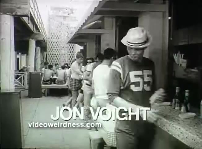 The still features Jon Voight prominently, wearing a sports jersey marked '55' and a straw hat. He appears to be in a casual outdoor setting, possibly at a diner or café, with patrons seated at tables in the background. The black-and-white image gives it a vintage feel that reflects the film's era, likely evoking themes of youth and social interactions. The text at the bottom clearly identifies him and includes a website reference.