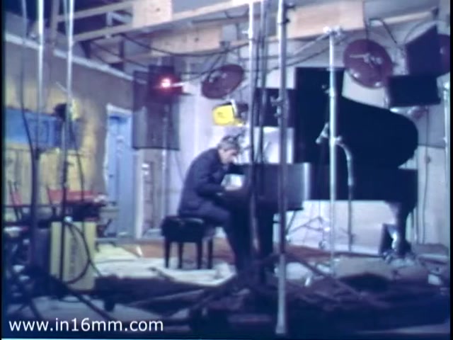 The still from the 'Burt Bacharach Christmas Seal PSA - 1975' features Burt Bacharach seated at a grand piano in a studio setting. The scene is illuminated by overhead lighting, and various microphones and equipment surround him, indicating a performance or recording session. The background has a casual, unfinished look, suggesting a staggered production environment. The overall atmosphere reflects a blend of creativity and seasonal spirit. Follow us on Twitter @FromFilm for more insights!