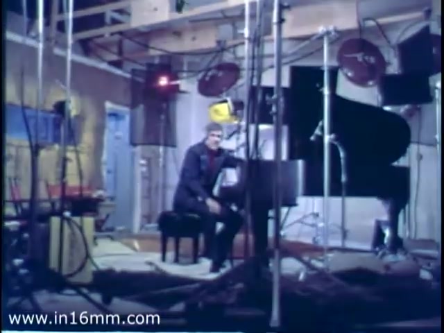 The still from the 'Burt Bacharach Christmas Seal PSA - 1975' features Burt Bacharach seated at a piano in a studio environment. Surrounded by various lighting equipment and sound gear, the scene conveys a candid moment in a recording setup. The studio appears somewhat unfinished, with exposed wood and a casual atmosphere, highlighting the behind-the-scenes nature of the production. Follow us on Twitter @FromFilm for more insights!
