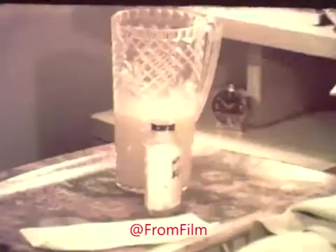 The still from 'Take Your Flu Shot Now! 1969' features a setting that emphasizes the importance of health and wellness. In the foreground, a small vial of what appears to be a vaccine sits on a tray. Behind it, there's a crystal pitcher containing a cloudy liquid, which could represent a health drink or remedy commonly associated with home care. The background is somewhat blurred, hinting at a domestic environment, likely a kitchen or an office space.   The overall composition reflects an era focused on public health and preventive measures, as indicated by the voice-over promoting vaccination against the flu. This still effectively captures the film's aim of encouraging viewers to take proactive steps in protecting their health during flu season.