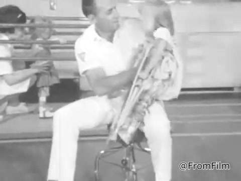 The still from the 'March of Dimes PSA - 1968' features a man in white attire, likely a sailor or uniformed personnel, sitting on a stool. He is playfully interacting with a young girl, who appears to be sitting on his lap, while holding a musical instrument. The background shows a casual setting, possibly a community event. The overall tone is light-hearted and emphasizes connection and joy between the adult and child.