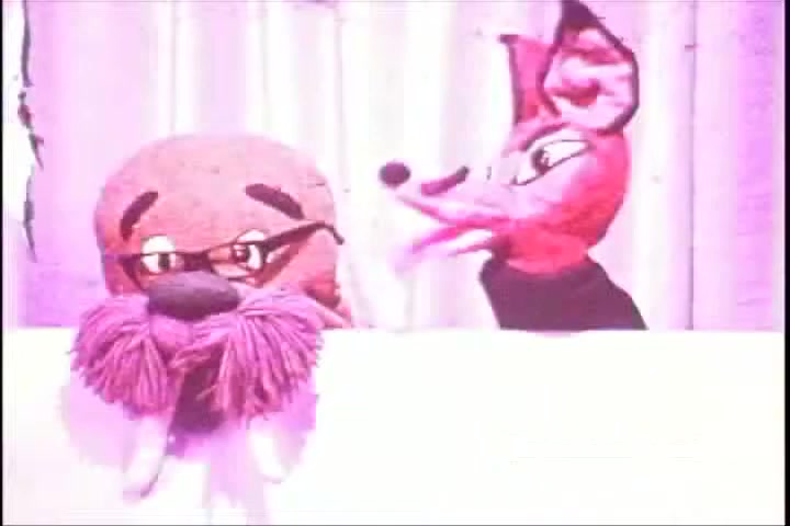 The still features two puppets: a walrus with glasses and a prominent mustache, and a vibrant, animated fox. The puppets are set against a backdrop of light-colored drapery, creating a whimsical and playful atmosphere typical of 1970s educational public service announcements. The walrus appears contemplative, while the fox is animatedly engaging, likely conveying a message about hearing. The overall color palette is muted with a soft pink hue, reflecting the era's aesthetic.