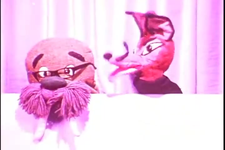 The still features two puppets, a walrus-like character with glasses and a bushy mustache, and a fox-like character with exaggerated features. They are positioned in front of a light pink background, likely part of a comedic or educational segment. The overall atmosphere suggests a playful approach, typical of public service announcements from the 1970s, aiming to engage viewers in a light-hearted manner while addressing hearing health.