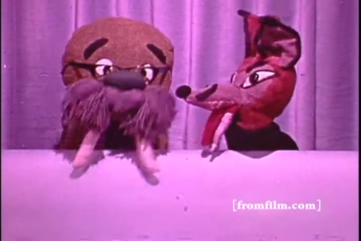 The still features two puppets, one resembling a walrus with glasses and a furry mustache, and the other resembling a cunning fox with exaggerated features. They are positioned against a backdrop of draped fabric, suggesting a stage setting. The vibrant colors typical of 1970s film are evident. The puppets appear to be engaged in conversation, likely conveying a message related to hearing awareness, consistent with the theme of the Better Hearing Institute PSA.