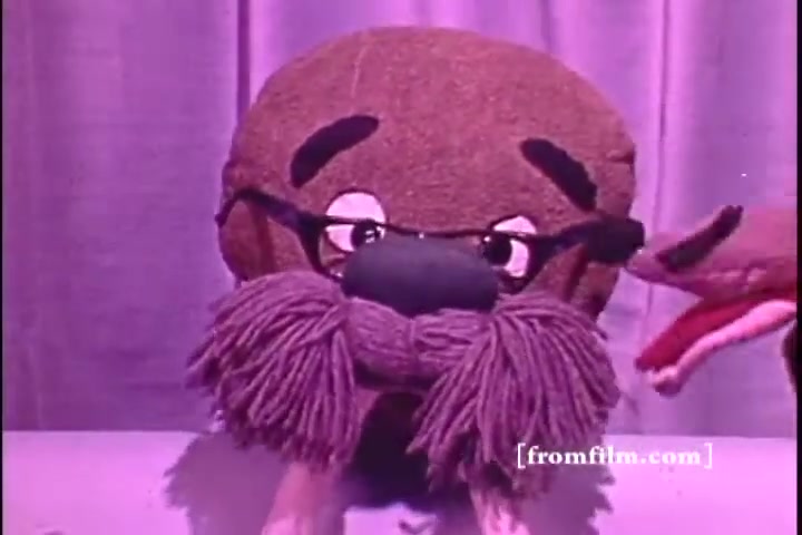 The still features a whimsical puppet character resembling a walrus, complete with glasses and a prominent mustache, set against a purple backdrop. Another puppet appears on the side, suggesting a playful interaction. The style and presentation reflect the educational and entertaining approach typical of 1970s public service announcements.