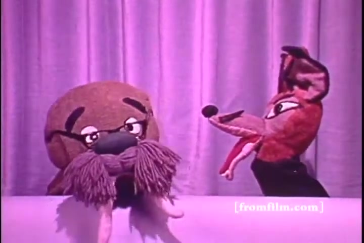The still features two puppets against a pastel backdrop. The left puppet resembles a walrus with large glasses and a bushy mustache, while the right puppet is a fox-like character with exaggerated features. Both are engaged in conversation, likely conveying a playful or educational message related to hearing awareness. The lighting is soft, evoking a vintage feel typical of 1970s public service announcements.