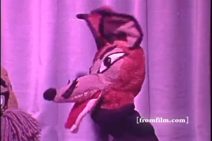 The still features a puppet resembling a fox, characterized by its exaggerated features and vibrant colors. The puppet has a prominent snout, large eyes, and intense expressions, likely conveying a message relevant to the theme of hearing awareness. The background is a soft purple curtain, creating a theatrical setting typical of a PSA from the 1970s. The overall feel is playful yet educational, aimed at engaging viewers on the topic of hearing health.