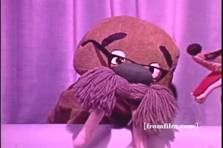 The still from the 'Better Hearing Institute PSA - 1970s' features a puppet character with a prominent mustache and glasses, appearing to be a whimsical figure. The background is a soft purple drape, setting a playful tone. Another puppet, possibly a dog or similar animal, is visible to the side, contributing to a lighthearted atmosphere. The overall aesthetics reflect the colorful and engaging style typical of public service announcements from that era.