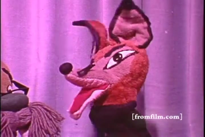 The still features a vibrant puppet with fox-like characteristics, including large ears and an expressive face. Its mouth is open as if speaking, creating a lively appearance. The background is a soft purple curtain, suggesting a stage setting typical of children's programming from the 1970s.