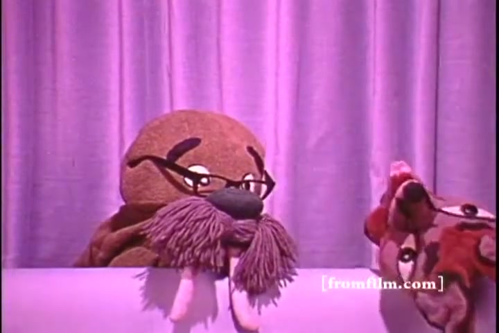 The still features two puppet characters in a setting reminiscent of a stage performance. One character, resembling a walrus with glasses and a large mustache, appears to be engaging with another puppet, possibly a playful animal. The backdrop is a soft purple curtain, creating a whimsical atmosphere. This scene likely conveys a lighthearted message related to hearing awareness, typical of the Better Hearing Institute PSA from the 1970s.