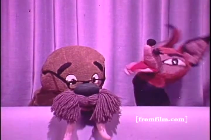 The still features two puppets against a backdrop of curtain drapes. The left puppet has glasses and a thick mustache, suggesting a wise or elderly character, while the right puppet is more vibrant with an animated expression. The whimsical scene likely aims to convey a message related to hearing awareness, typical of public service announcements from the 1970s. The muted colors and vintage style reflect the period's aesthetic.