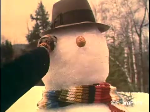 The still from the 'Diamond Nuts Commercial - 1970s?' showcases a snowy outdoor setting where a snowman is being adorned. A hand reaches out to place a piece, likely a nut, as the snowman sports a hat and a colorful scarf. The warm, nostalgic tones evoke a simple, cheerful winter scene, typical of commercials from that era. Follow for more film insights @FromFilm.