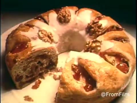 This still from the 1970s Diamond Nuts commercial features a close-up of a beautifully arranged baked good, likely a bread or cake adorned with nuts and cherries. The texture appears soft, with a glossy icing on top, enhancing its visual appeal. The composition captures the warm, inviting essence of home-baked treats, typical of nostalgic food advertising from that era. The vibrant colors and rich details draw attention to the nuts embedded in the dessert, emphasizing their role in the product.   Follow us on Twitter @FromFilm.