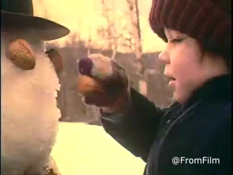 The still from the 1970s Diamond Nuts commercial features a snowy outdoor setting. A child with a burgundy knit cap interacts with a snowman, which is adorned with nut accessories. The child appears focused as they reach out with a gloved hand. The warm color palette evokes a nostalgic winter atmosphere. This scene captures a playful moment highlighting the theme of joy associated with the product. Follow us on Twitter @FromFilm.