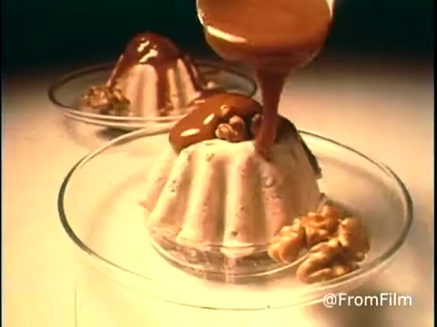 The still from the 'Diamond Nuts Commercial - 1970s' showcases two elegant dessert molds as a thick, glossy sauce is poured over one, highlighting the texture and richness. Walnuts are placed attractively around the dessert, emphasizing the nutty theme of the brand. The lighting gives a warm, inviting feel typical of advertisements from that era, focusing on indulgence and pleasure. The overall composition is visually appealing, capturing the essence of a classic food commercial.