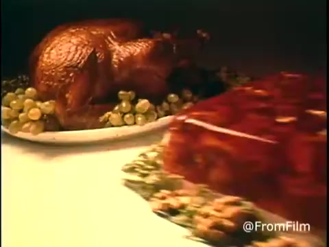 The still from the 'Diamond Nuts Commercial - 1970s' features a beautifully presented table with a roasted turkey and a glossy jelly dessert. The turkey is garnished with green grapes, creating a festive and appetizing scene typical of 1970s holiday advertising. The colors are warm and inviting, emphasizing the home-cooked feel of the meal. The composition suggests a focus on traditional family gatherings and the role of Diamond Nuts in those culinary moments.