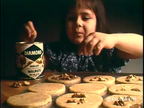 The still from the 'Diamond Nuts Commercial - 1970s' features a young girl focused on preparing cookies, with a can of Diamond Shelled Walnuts prominently displayed. She’s sprinkling chopped nuts onto cookie dough, epitomizing a homey and familial feel typical of the era's advertising. The warm colors and inviting atmosphere capture the essence of 1970s kitchen culture.