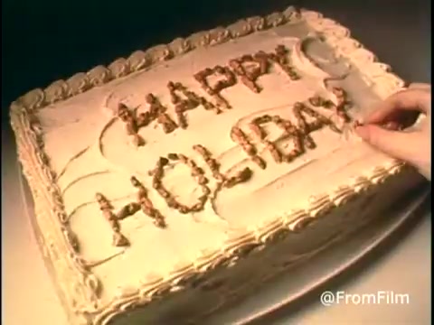 The still from the 'Diamond Nuts Commercial - 1970s' features a close-up of a cake, adorned with cream frosting and decorated with the words 'HAPPY HOLIDAY' spelled out in nuts. A hand is seen carefully placing the nuts to form the letters, highlighting the brand's nut product. The overall ambiance is festive and inviting, capturing the spirit of celebration typical of holiday advertisements from that era. Follow us on Twitter @FromFilm.