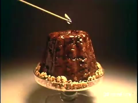 The still from the 'Diamond Nuts Commercial - 1970s' features a glossy, chocolate-covered dessert displayed on a glass pedestal. A skewer pierces the top of the cake, suggesting a focus on texture and indulgence. The dessert is surrounded by what appears to be scattered nuts, emphasizing the product being advertised. The lighting enhances the rich colors, creating an inviting visual appeal, typical of commercial aesthetics from that era.