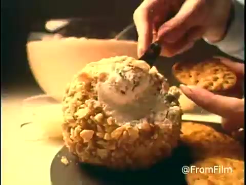 The still from the 'Diamond Nuts Commercial' depicts a hand scooping out of a large, round cheese ball coated in nuts. Next to it, there are chocolate chip cookies, creating a tempting visual snack combination. The lighting adds warmth, catering to a nostalgic feel typical of 1970s commercials.