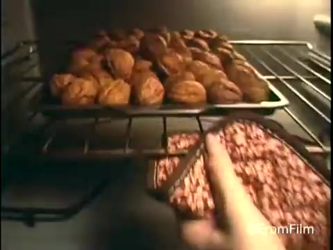 The still from the 'Diamond Nuts Commercial - 1970s' features an oven scene focusing on a baking tray filled with freshly roasted nuts. The tray's contents include both walnuts and possibly another nut variety, showcasing a homey, nostalgic kitchen setting typical of advertisements from that era. A hand is reaching toward the tray, suggesting preparation or serving, which emphasizes the product's appeal. The warm tones and the framing create an inviting atmosphere, aligning with the commercial's intent to promote Diamond Nuts as a wholesome, delicious snack.   Follow us on Twitter @FromFilm.