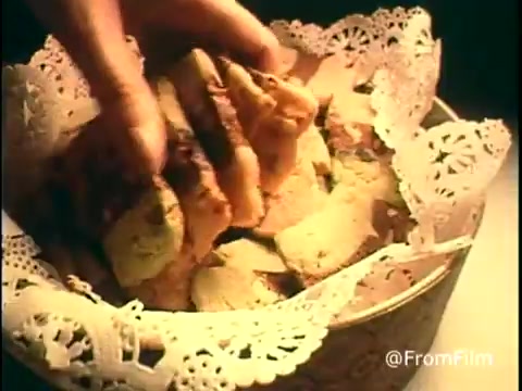 The still from the 'Diamond Nuts Commercial - 1970s' features a hand reaching into a decorative bowl lined with lace-like paper. The bowl appears to contain a mix of nuts and possibly other snack items, presenting a warm and inviting look typical of 1970s advertising. The overall aesthetic invokes a cozy, nostalgic feel associated with home and gatherings.
