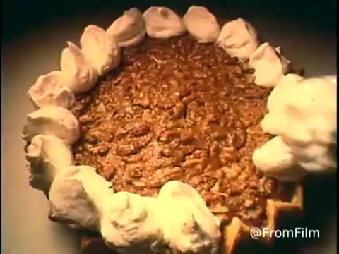 The still from the 1970s Diamond Nuts commercial features a close-up of a pecan pie topped with generous swirls of whipped cream. The pie's surface is rich with pecans, showcasing its delectable appeal. The image highlights the texture and colors of the pie, evoking a sense of warmth and home baking. Follow us on Twitter @FromFilm for more nostalgic content!