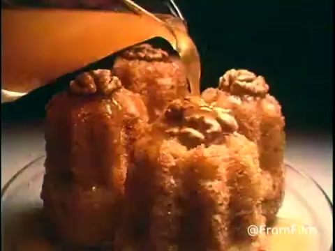 The still from the 'Diamond Nuts Commercial - 1970s' features a close-up shot of fluffy, bundt-shaped desserts topped with walnuts. A golden syrup is being poured over them, emphasizing a rich, inviting visual appeal. The lighting highlights the textures and sheen of the desserts, creating an appetizing scene that showcases the product prominently.