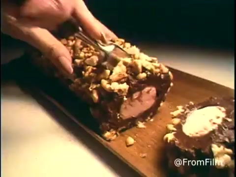 The still from the 'Diamond Nuts Commercial - 1970s' features a close-up of a hand using a knife to slice through a chocolate-covered log. The treat is adorned with crushed nuts, creating an appealing texture. The background is simple, emphasizing the product, likely designed to highlight indulgence and deliciousness. The retro aesthetic of the 16mm print adds to the nostalgic vibe of the advertisement.