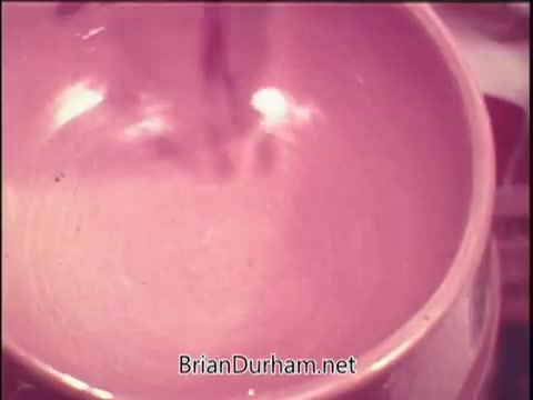 The still from the Gaines Meal dog food commercial features a close-up of a mixing bowl. It captures the action of stirring, likely showcasing the preparation process of the dog food. The color palette has a warm, nostalgic hue typical of 1970s advertising. The overall focus appears to be on the texture and appeal of the food being prepared for dogs.