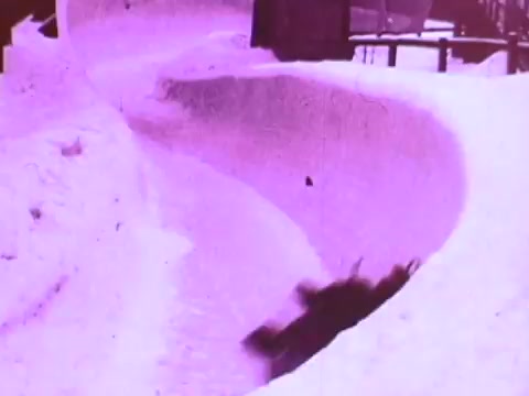The still features a snowy, curved bobsled or luge track surrounded by high snowbanks. In the foreground, a figure is seen gliding down the track, suggesting motion and speed, likely emphasizing the performance and reliability of Mobil I oil in extreme conditions. The overall hue of the image is a distinct purple, lending a vintage feel typical of 16mm film.