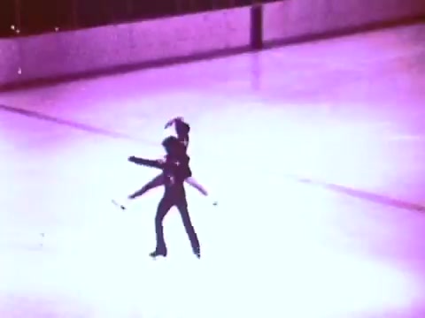 The still features two figure skaters performing a synchronized routine on ice, captured in a vintage 16mm style. The scene is lit with a soft, purplish hue, highlighting their movement and grace as they glide across the rink, embodying elegance and fluidity. The backdrop is a blurred ice surface, emphasizing their dynamic motion.